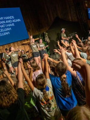 Summer staff leading worship from stage, campers raising hands in worship at Deer Run Camps & Retreats summer camp.