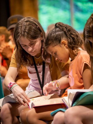 Female camp counselor helps her girl camper find a reference in her bible during the nightly sermon at summer camp.