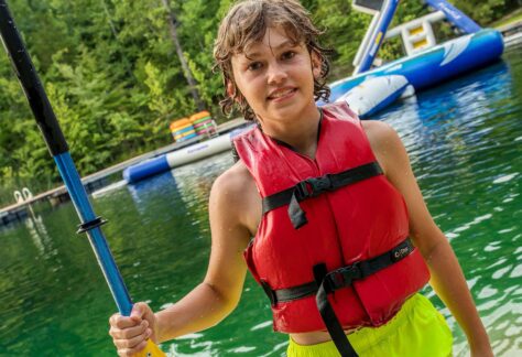 overnight summer camp boy holding corcl boat paddle wearing red life jacket with aqua park in background at Deer Run Camps & retreats